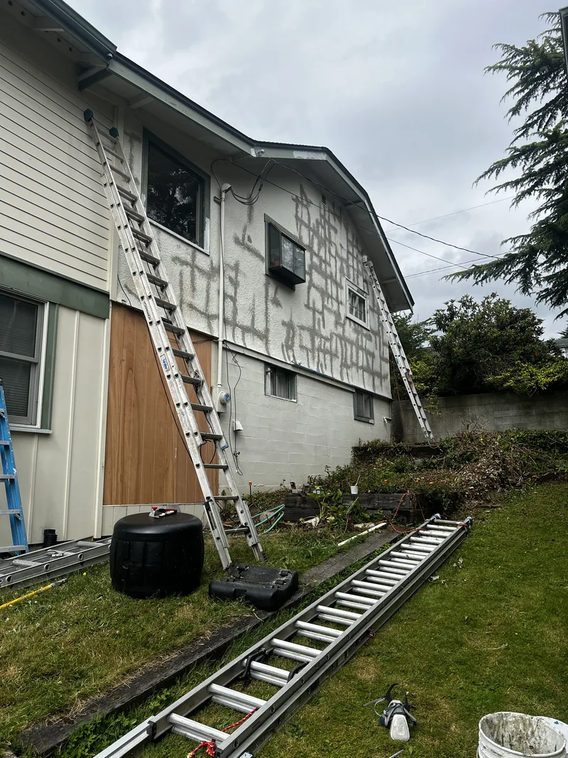 Stucco cracks cut out and patched across the upper wall of a Fairhaven home in Bellingham, WA, with ladders set for the repair.